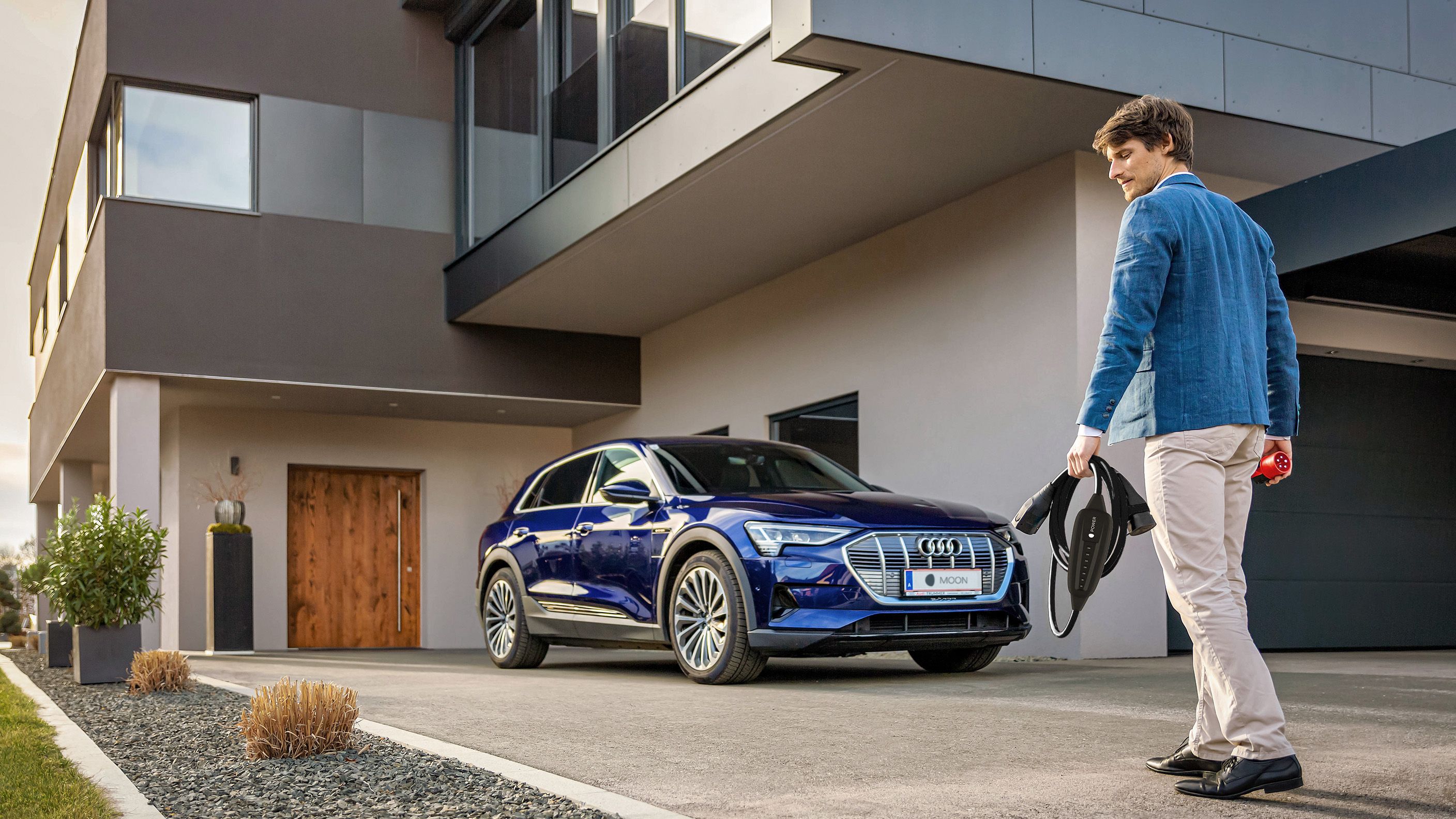 Mann geht mit einem Kabel in der Hand auf ein E-Auto bei einem Haus zu